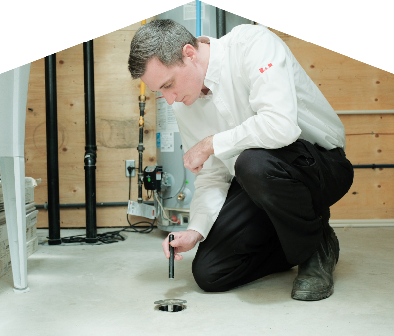 Home Aire Care plumbing specialist inspecting a basement floor drain with a flashlight during a drain cleaning service, in a utility room with visible pipes, water heater, and wood panel walls.