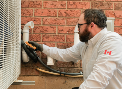 A Home Aire Care HVAC specialist wearing glasses and gloves uses a diagnostic tool to service an outdoor AC unit beside a brick wall. A Home Aire Care HVAC specialist wearing glasses and gloves uses a diagnostic tool to service an outdoor AC unit beside a brick wall.